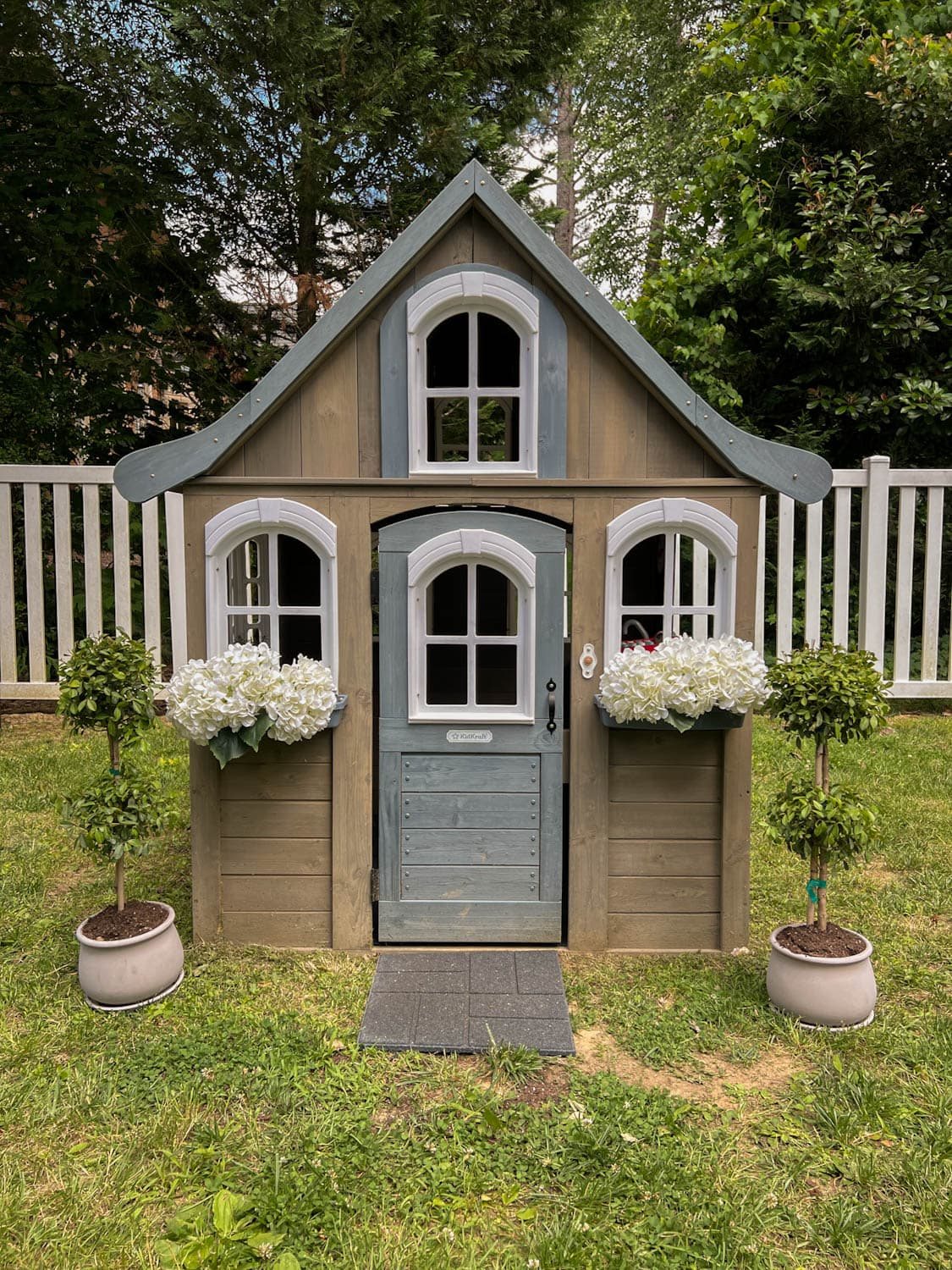 wooden outdoor playhouse with white flowers and topiaries