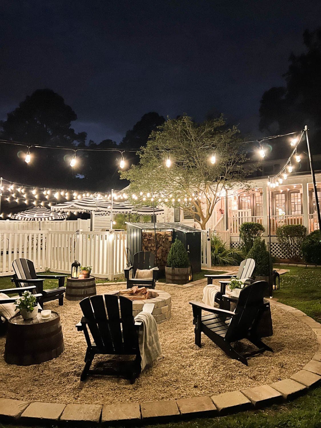 fire pit string lights with pea gravel, black Adirondack chairs, and wood shed in a backyard
