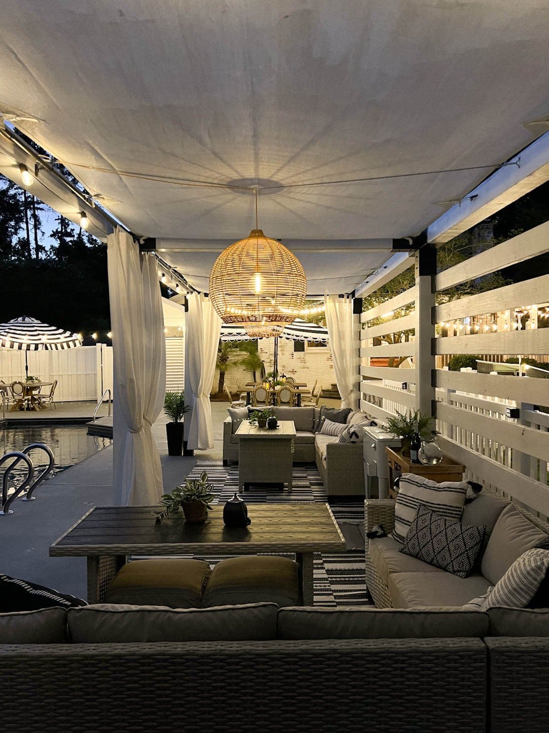 a pergola cabana with patio dining furniture, solar pendant lights, and string lights beside a pool at night