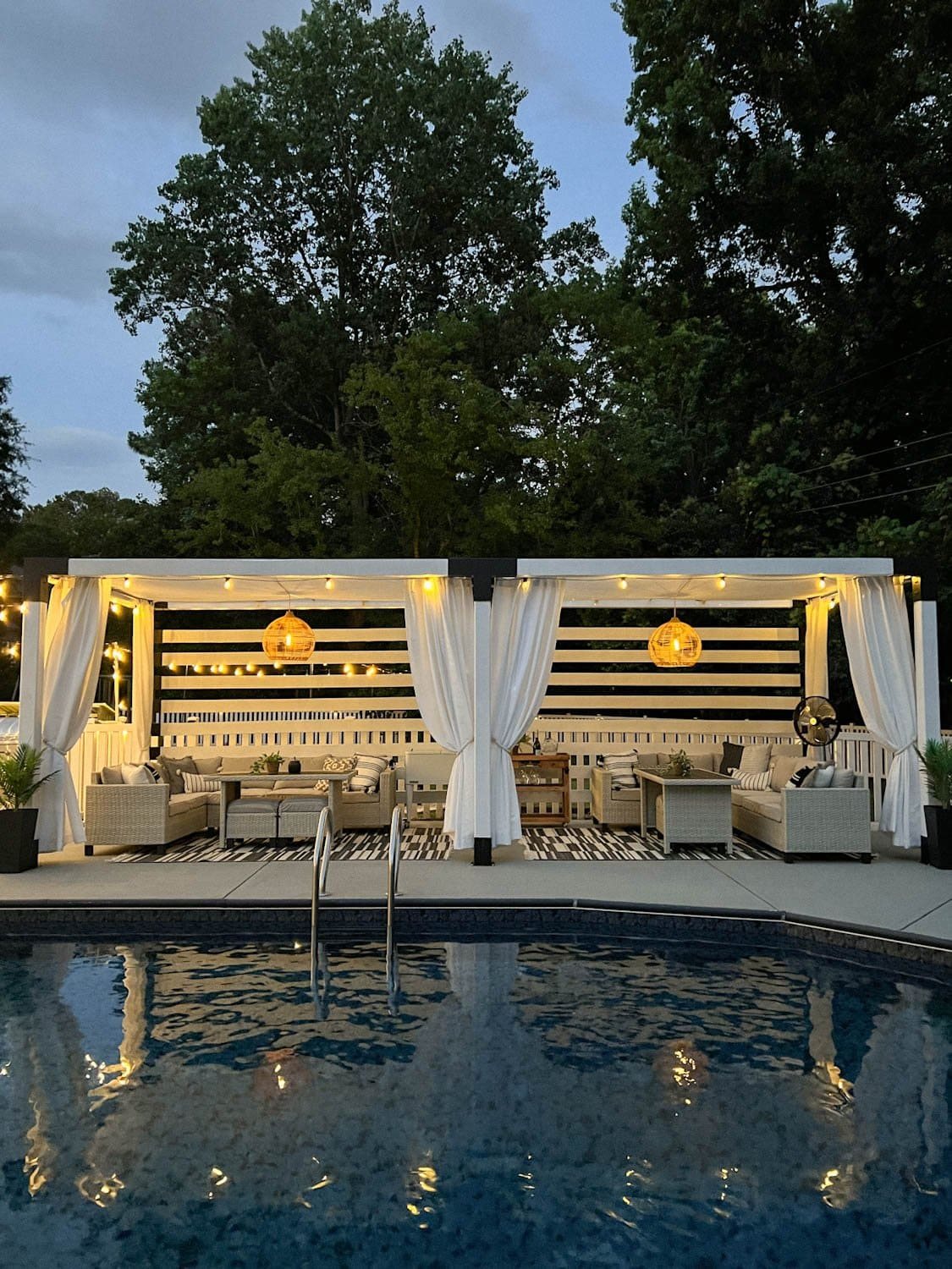 poolside cabana pergola in a backyard with string lights and solar pendant lights and white outdoor curtains