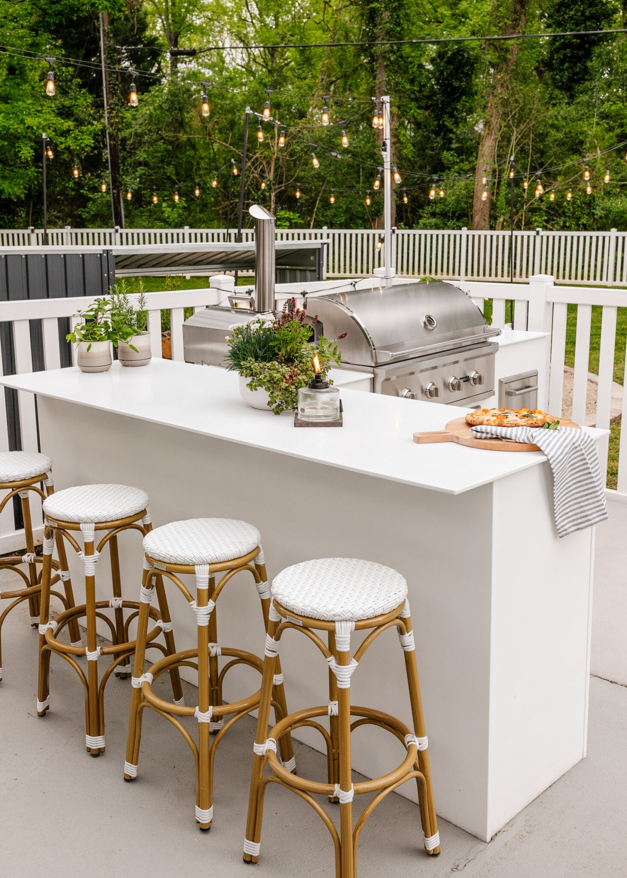 white outdoor kitchen with grill, bar stools, string lights, white fence, and potted plants