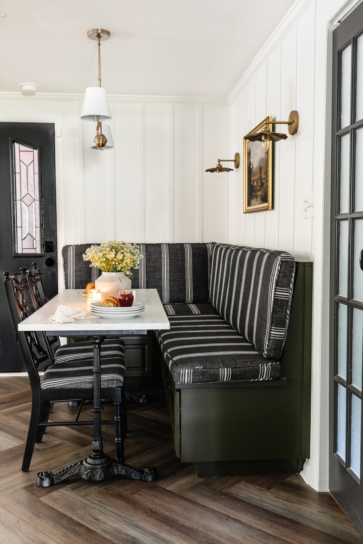 cottagecore breakfast nook with banquette, shaded chandelier, bifold French door, herringbone floor, and marble bistro table
