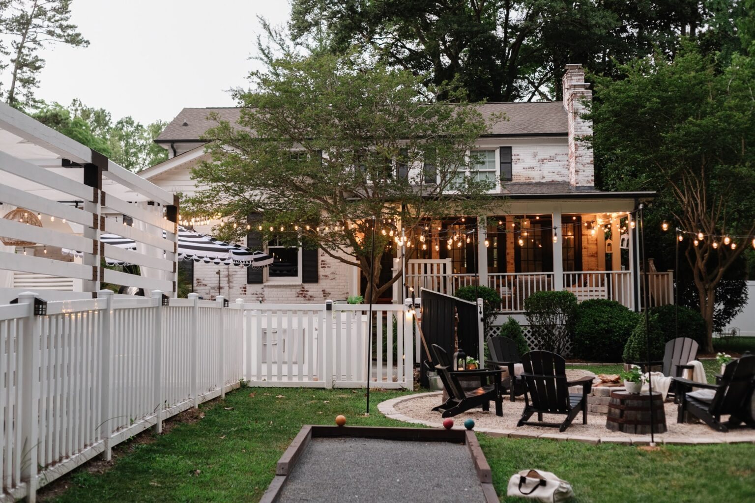 limewashed brick house, backyard fire pit, bocce ball court, white fence, back porch, and string lights
