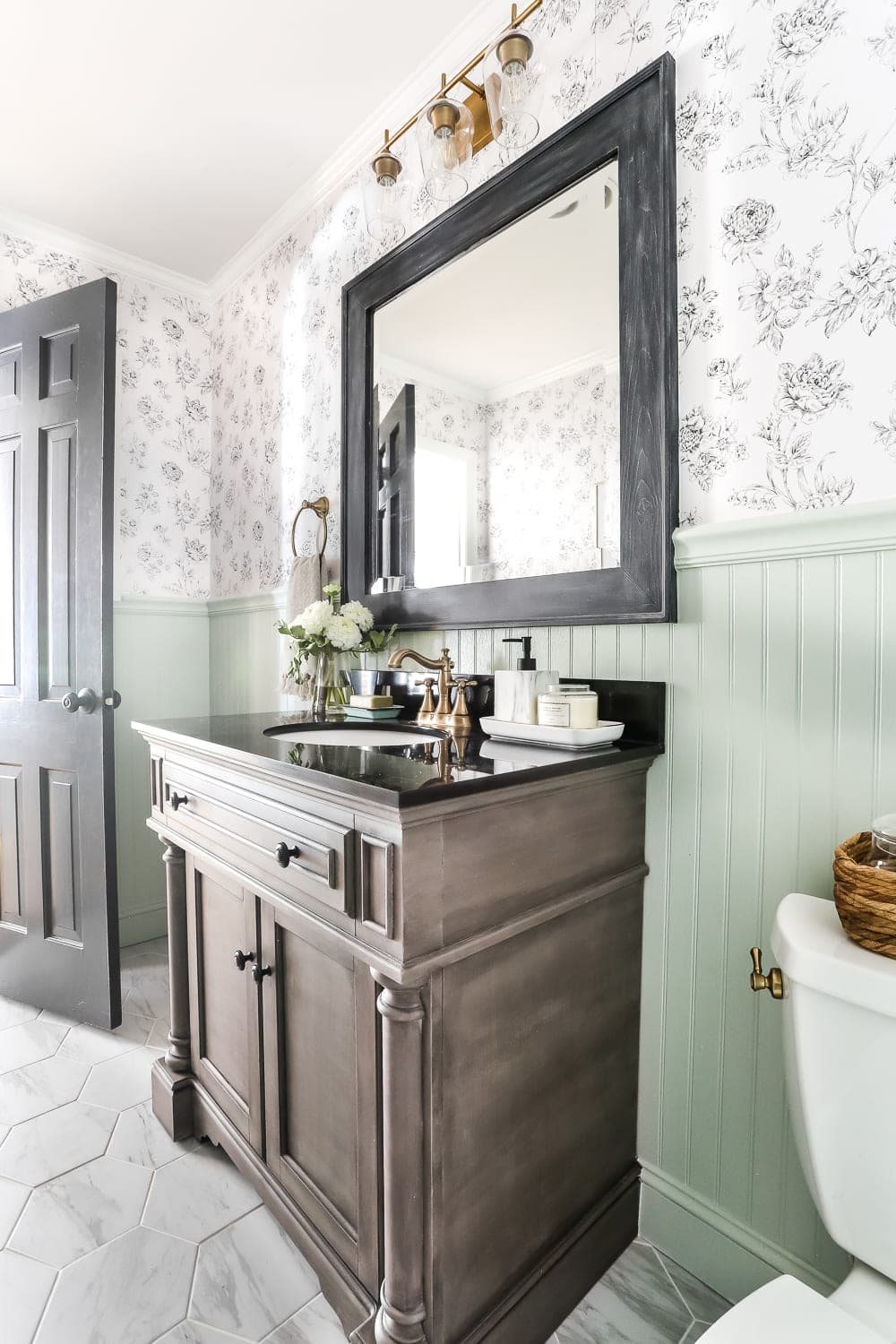 mint green beadboard in a half bath with black and white floral wallpaper, faux marble hexagon tile floor, and wood vanity