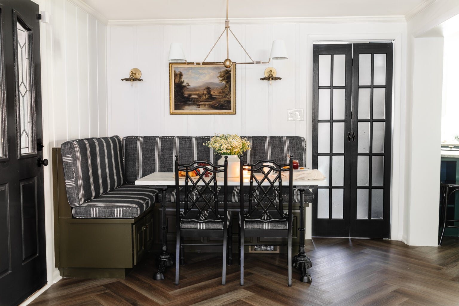 cottagecore breakfast nook with banquette, shaded chandelier, bifold French door, herringbone floor, and marble bistro table