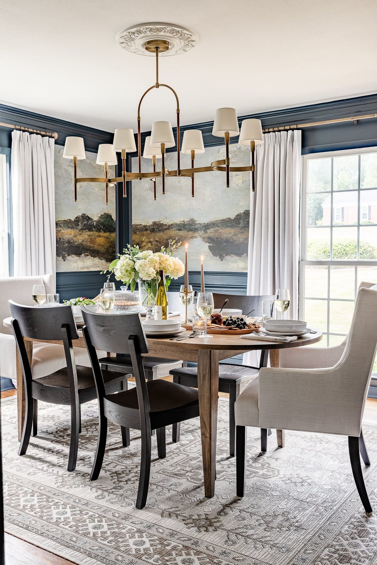 navy blue dining room with mural walls in picture frame molding, linen curtains, shaded chandelier, wood oval dining table, linen head chairs, and black dining chairs