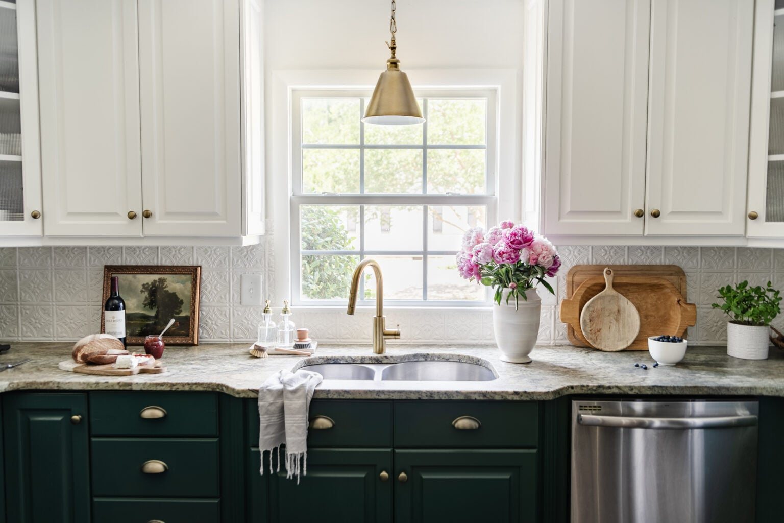 kitchen sink in front of window with white vase of pink peonies, antique art, and cuttingboards on a countertop