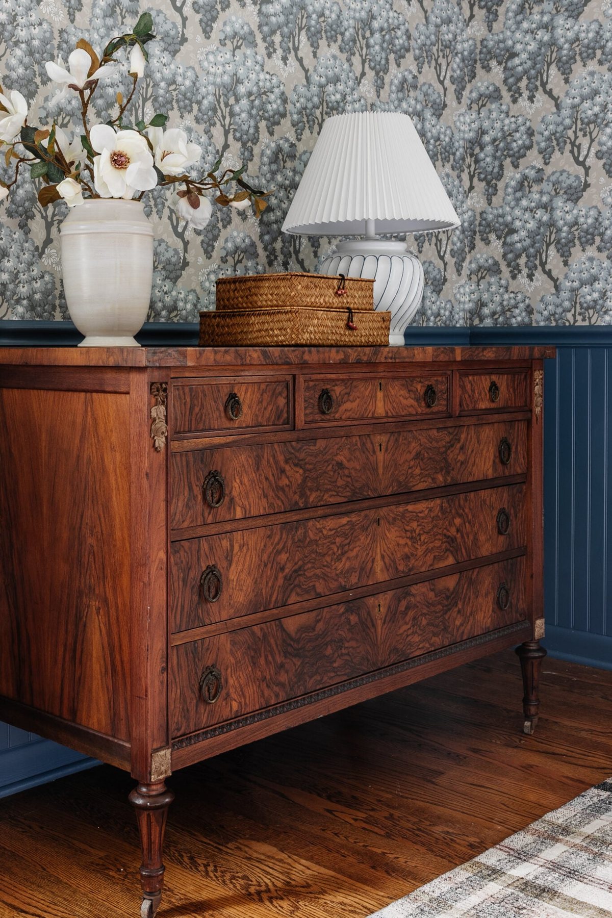burl wood dresser styled with vase of flowers woven basket boxes and lamp in a bedroom with blue vintage wallpaper