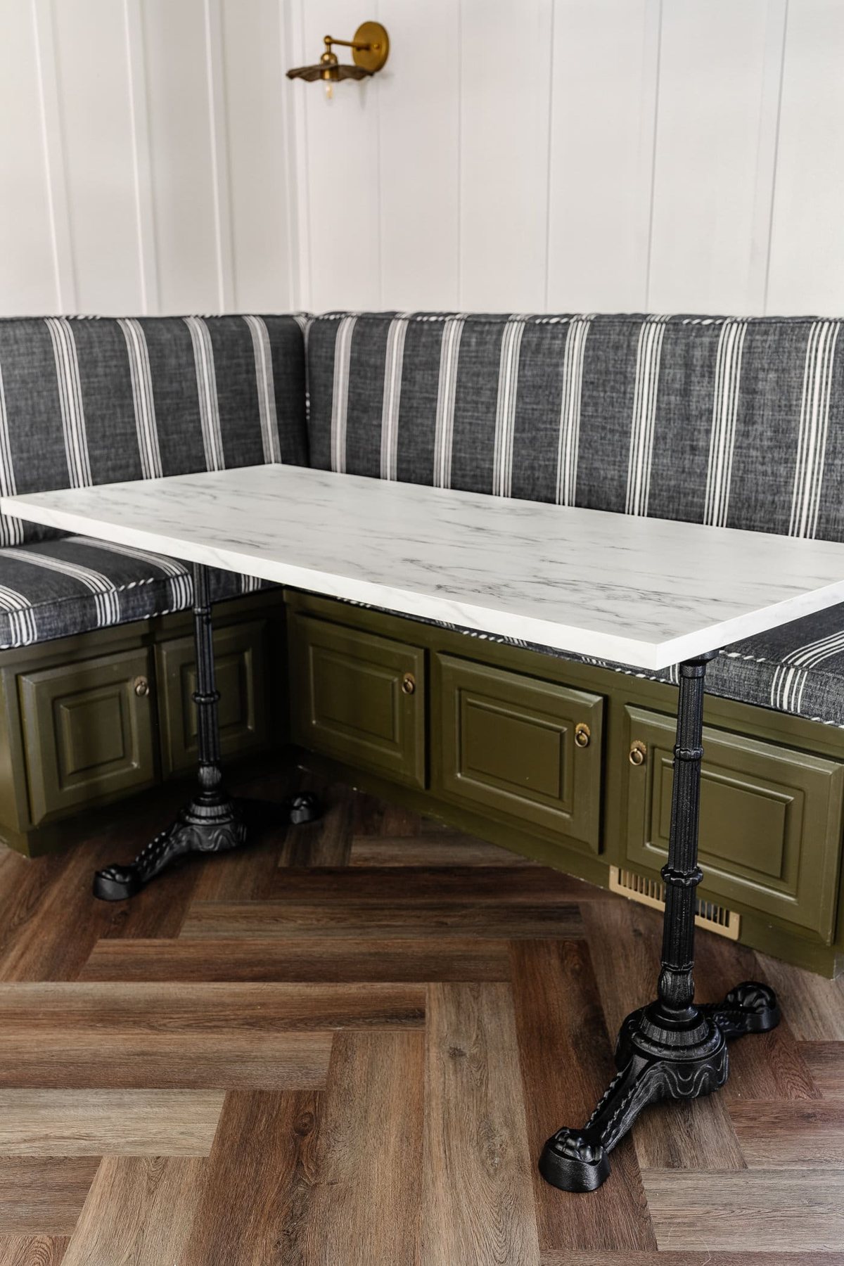 Parisian style faux marble dining table with cast iron pedestal legs against an olive green banquette with charcoal striped bench cushions and herringbone wood floors