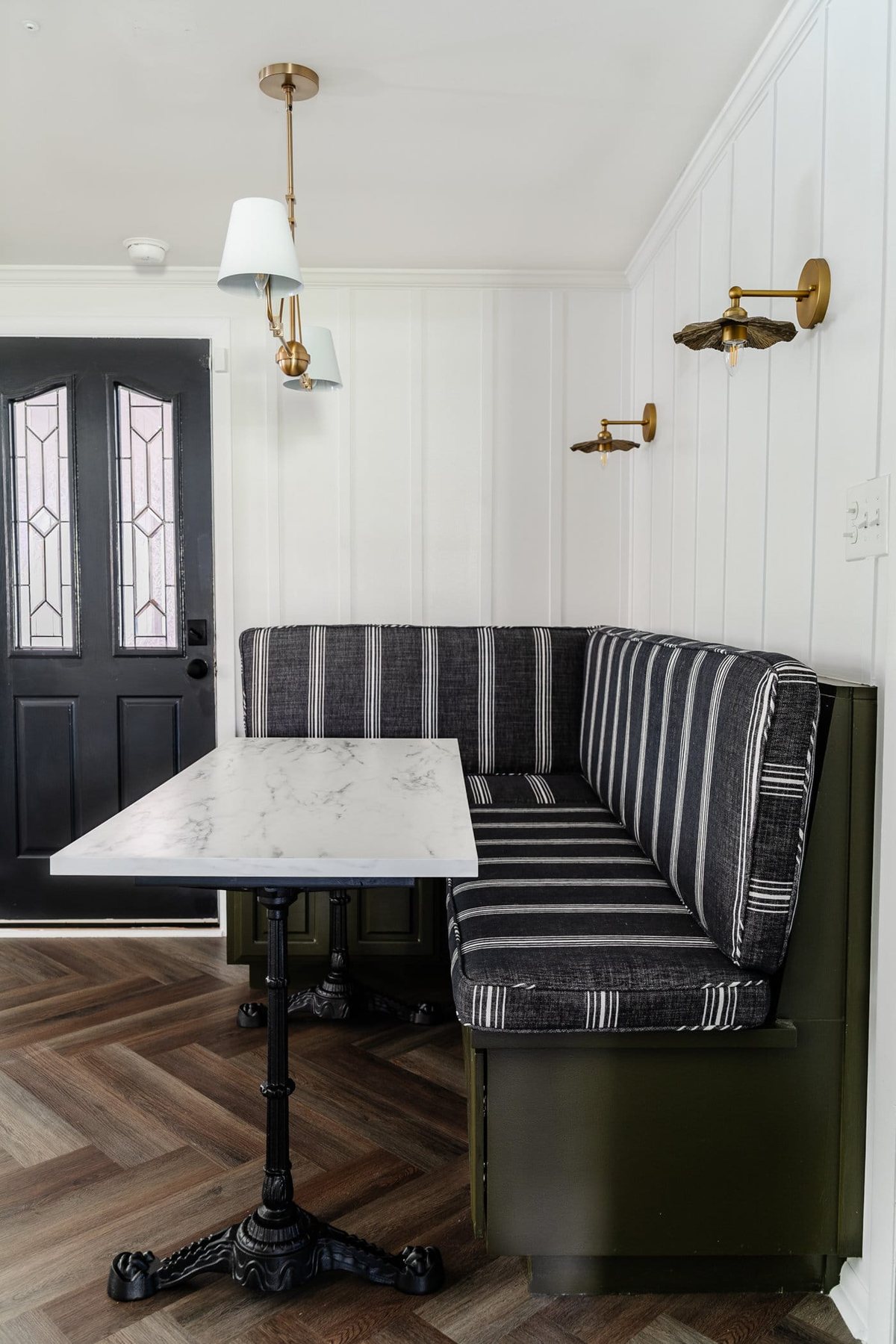 Parisian style faux marble dining table with cast iron pedestal legs against an olive green banquette with charcoal striped bench cushions and herringbone wood floors
