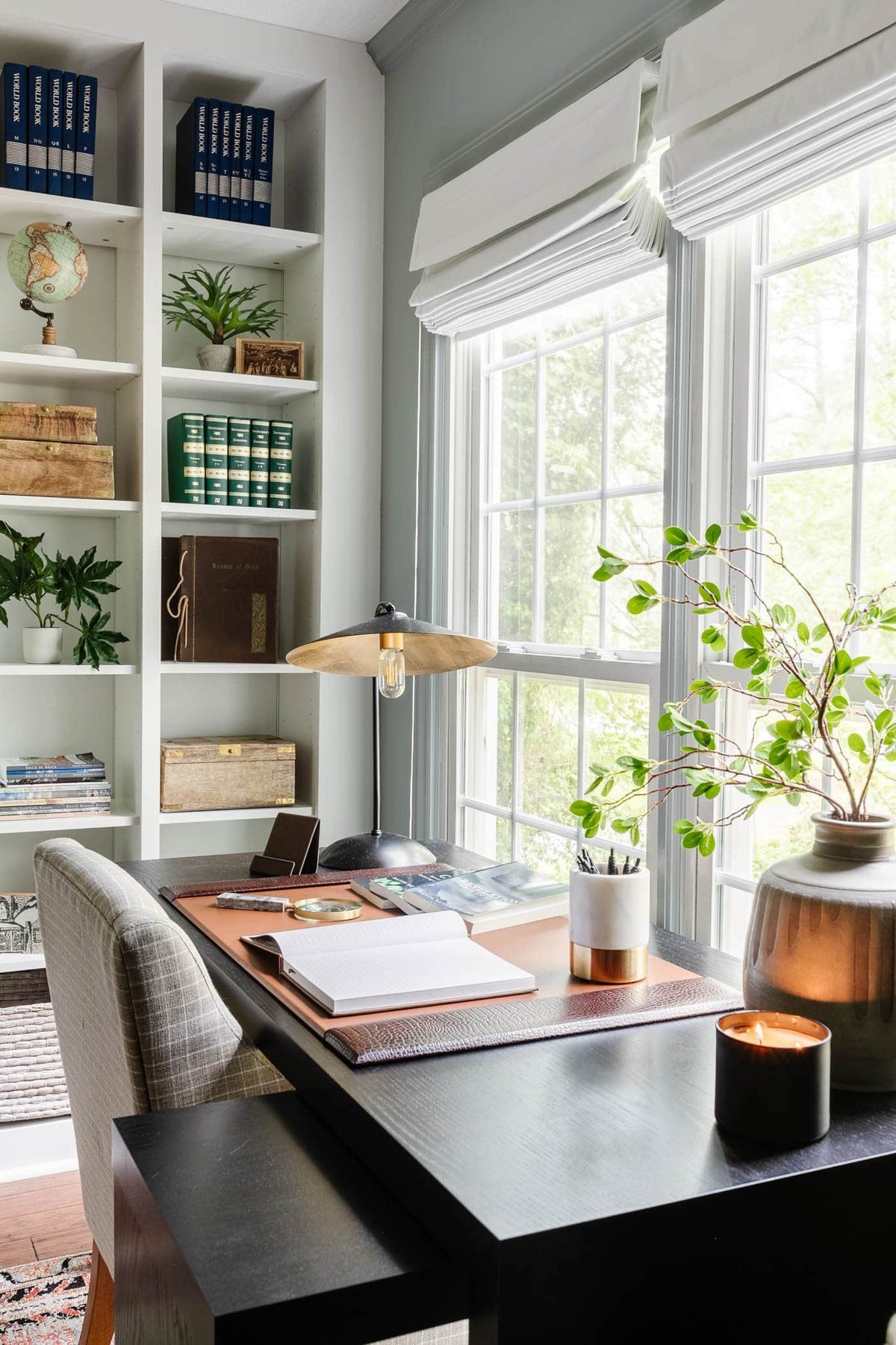black office desk in front of window with desk pad, desk lamp, vase of branches, and shelving