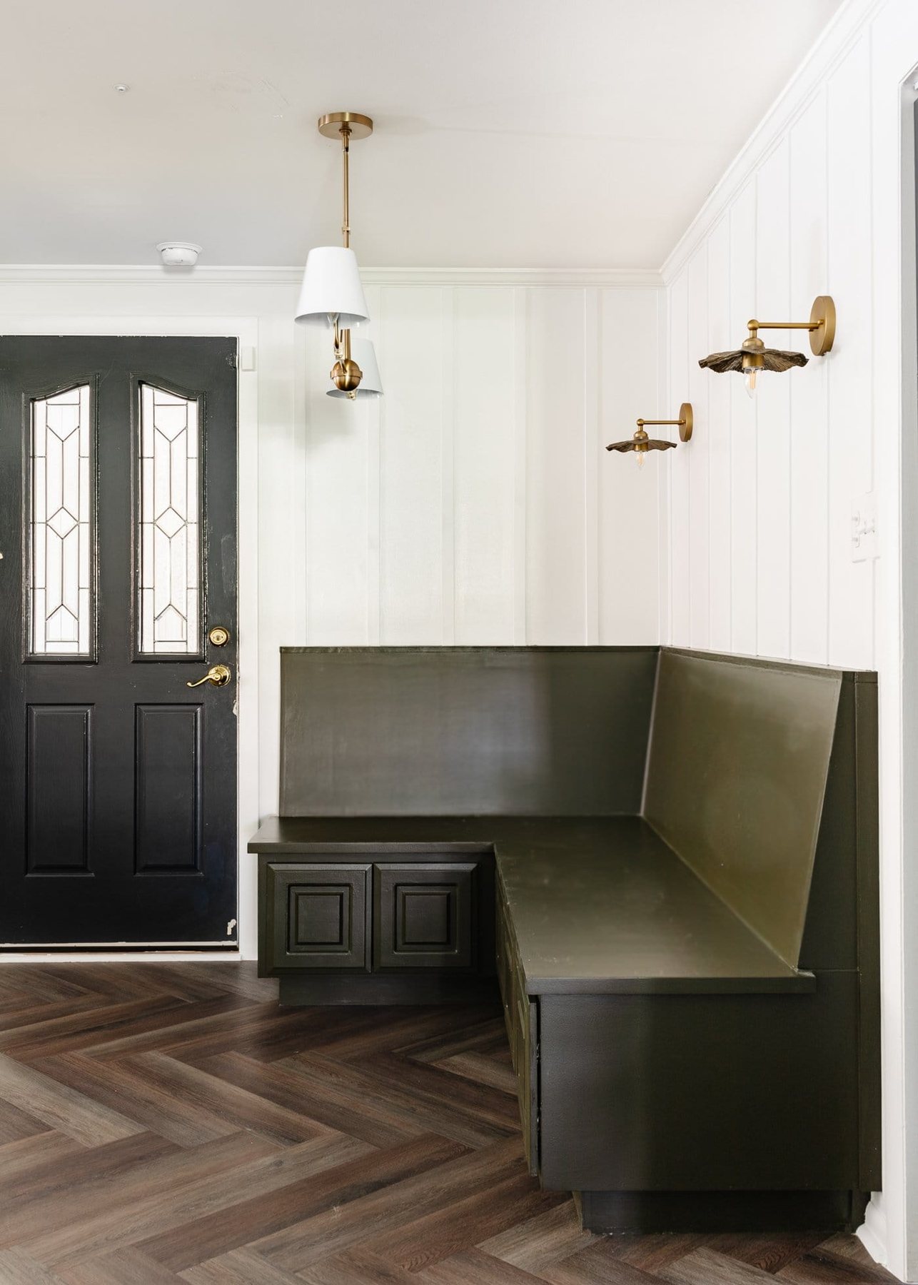 olive green banquette bench in a breakfast nook with white batten, herringbone floors, and black door