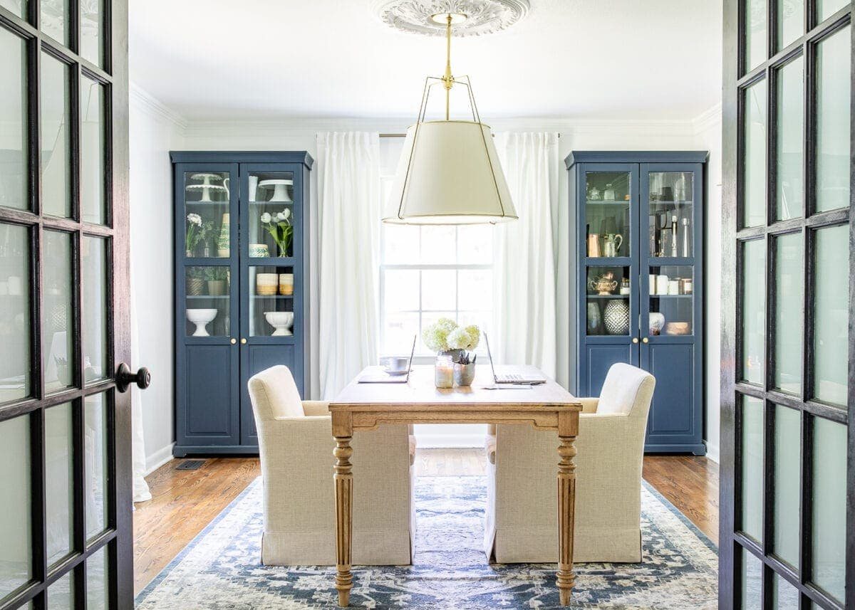 home office for two with work table and rolling arm chairs, white walls, navy blue storage cabinets, and black French doors