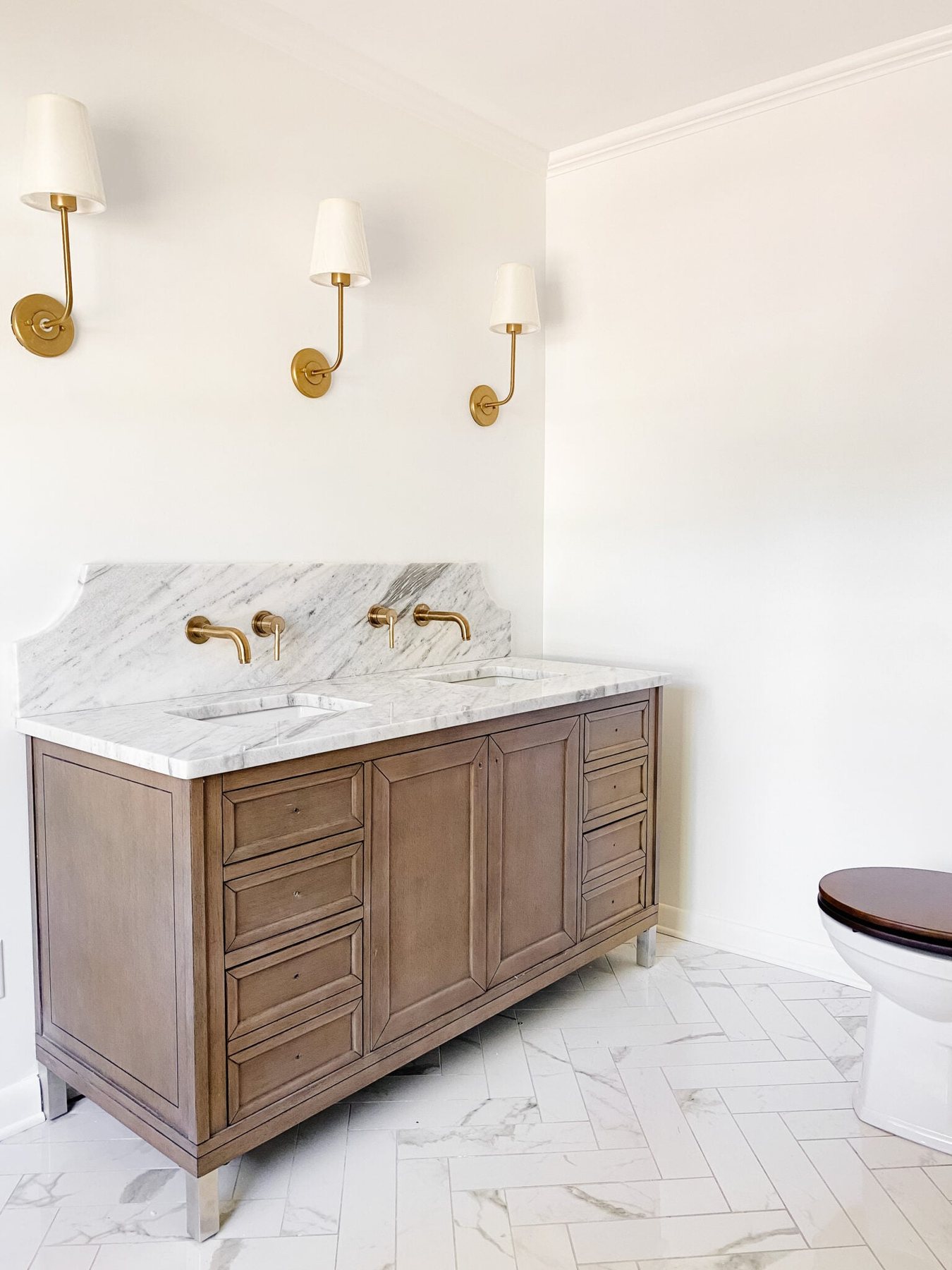 scalloped marble backsplash with gold wall mount faucets, faux marble porcelain tile herringbone floor, and wooden vanity in en suite bathroom