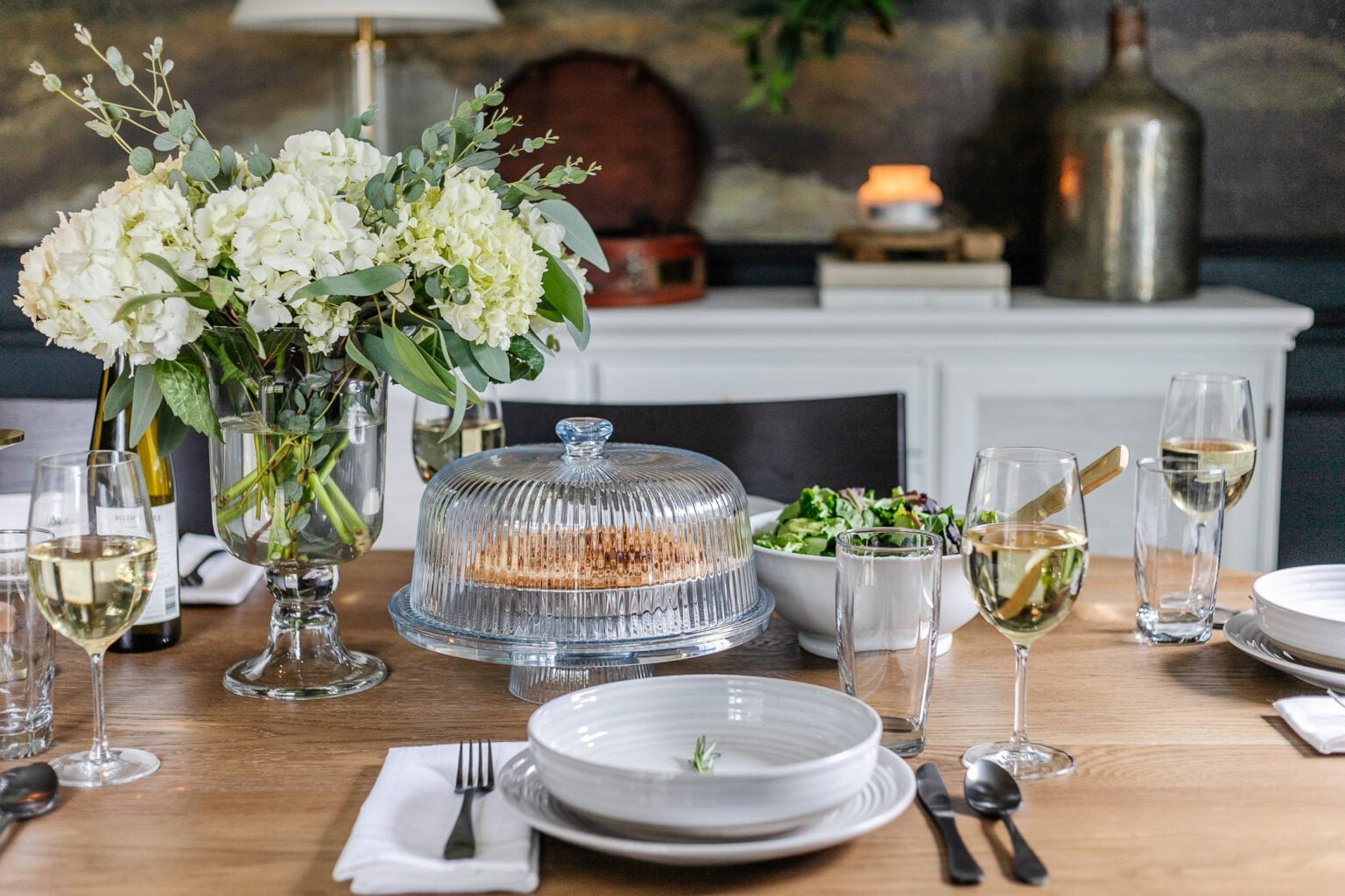 vase of hydrangeas, simple place setting of gray dishes and sprig of rosemary, cake dome, and serving bowl of salad
