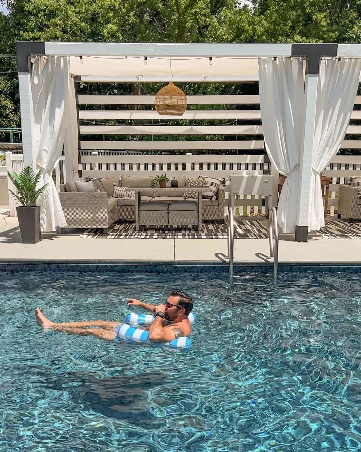 man floating on a hammock pool float in a swimming pool in front of a cabana