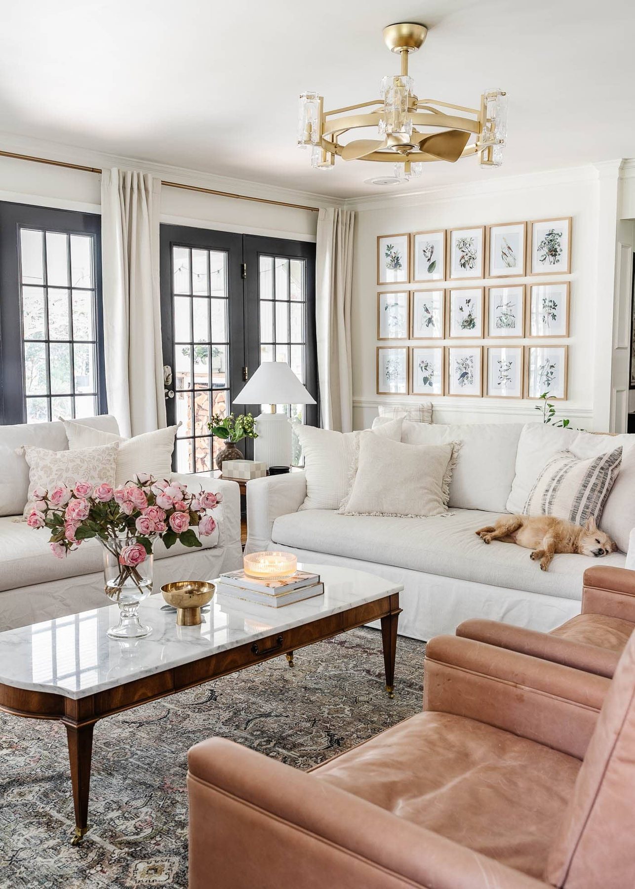 white living room with limewashed brick fireplace, gold fandelier, white Pottery Barn sofas, leather recliners, art TV, marble coffee table, and black French doors