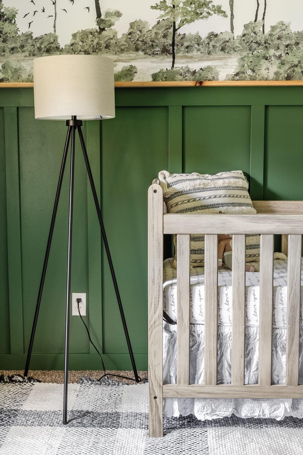 woodland theme baby boy nursery with floor lamp, natural wood crib, plaid rug, and wall mural