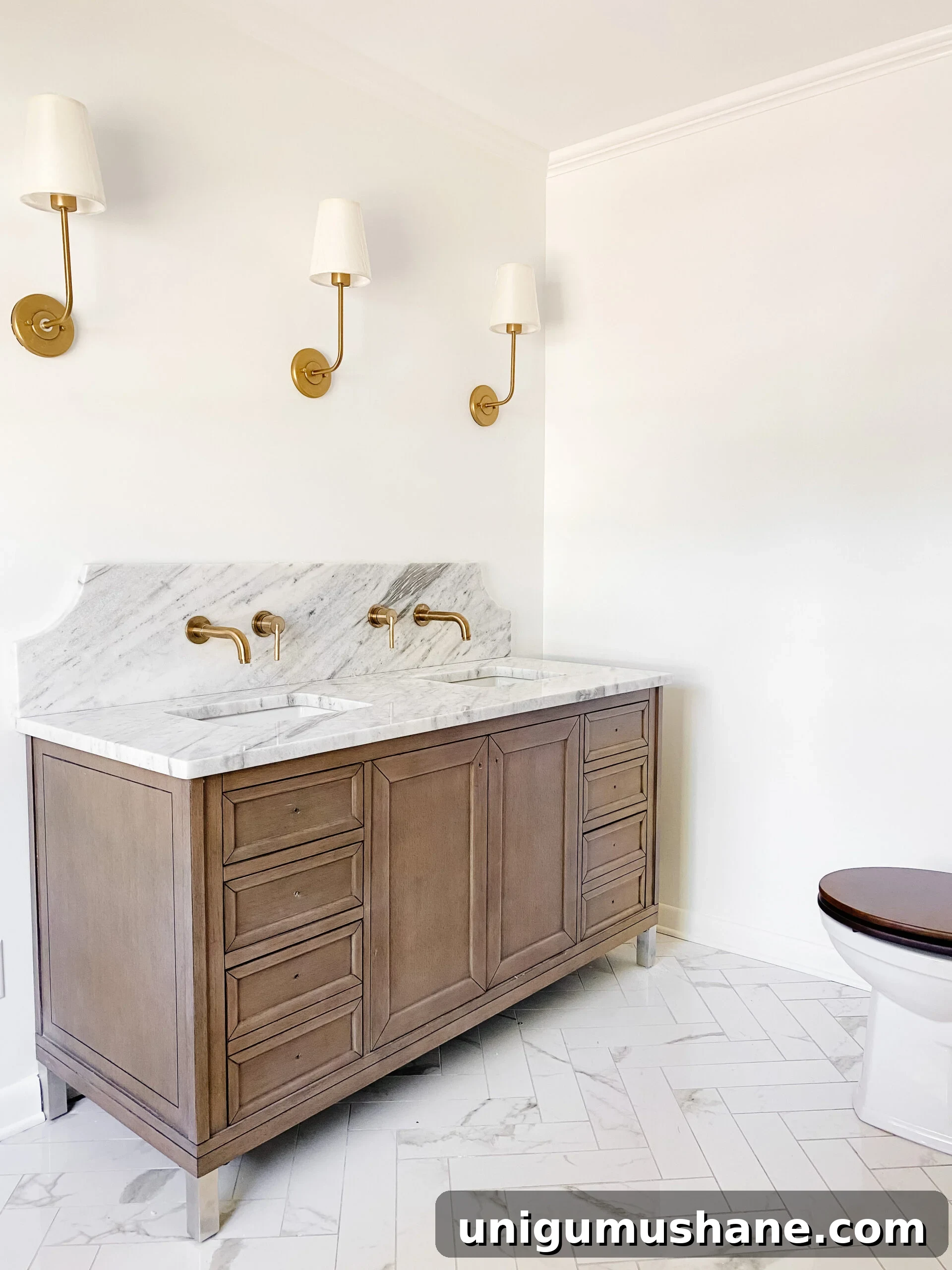 En Suite Bathroom Remodel: New Faucets Installed 2 scalloped marble backsplash with gold wall mount faucets, faux marble porcelain tile herringbone floor, and wooden vanity in en suite bathroom
