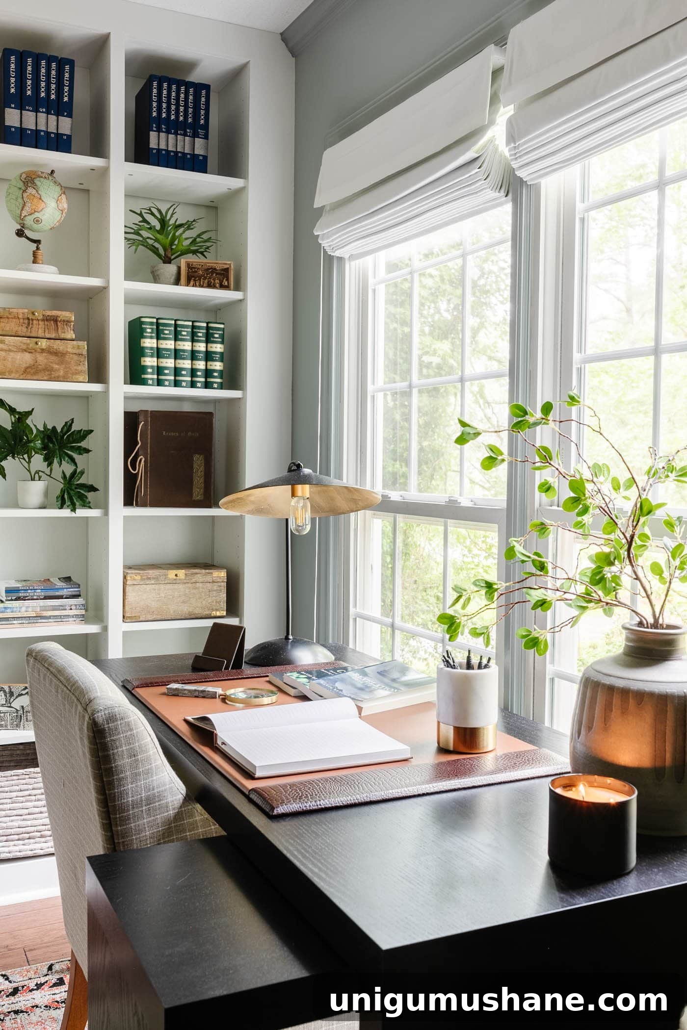 black office desk in front of window with desk pad, desk lamp, vase of branches, and shelving