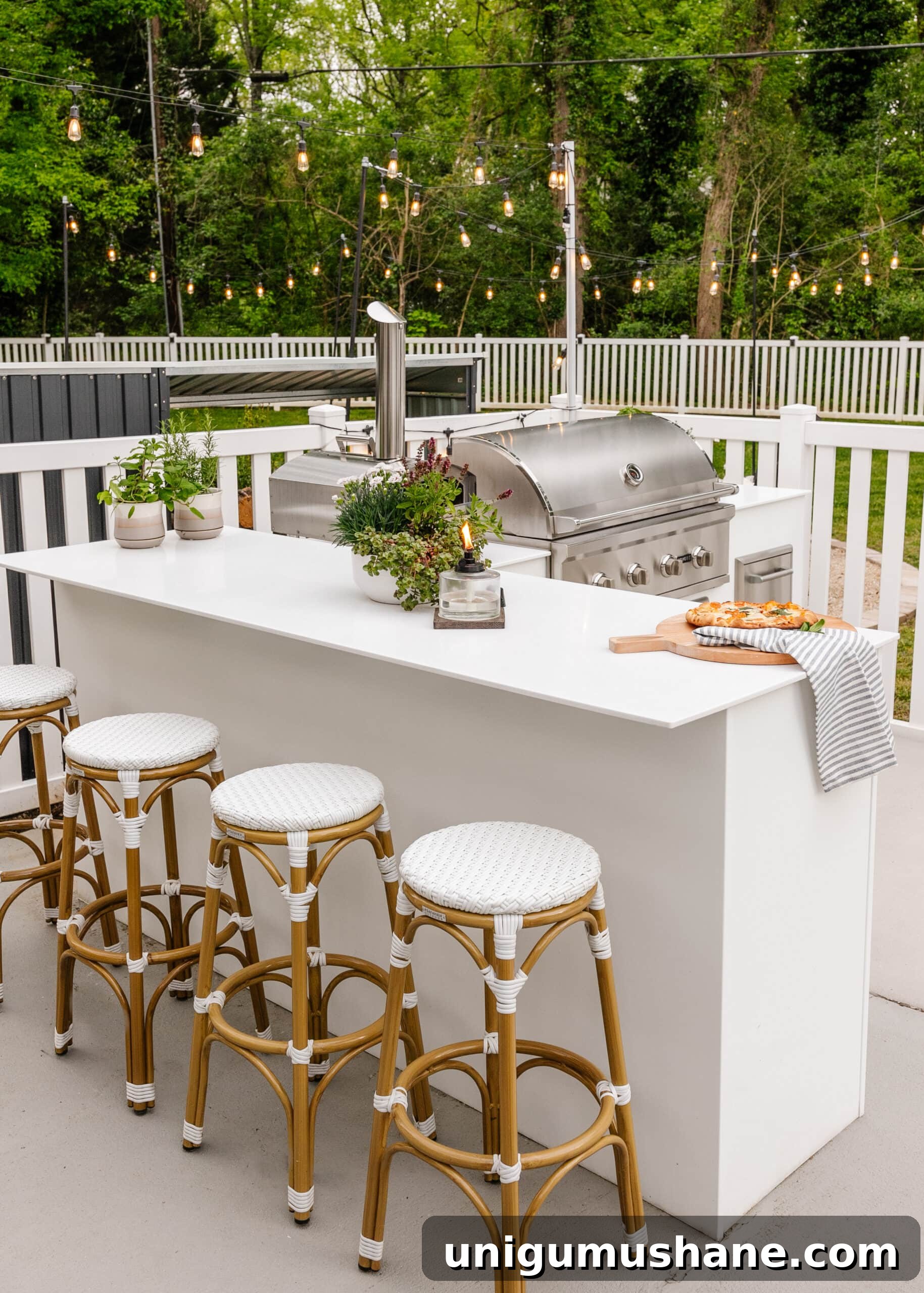 Spring Cleaning Checklist for Your Backyard Patio 8 white outdoor kitchen with grill, bar stools, string lights, white fence, and potted plants